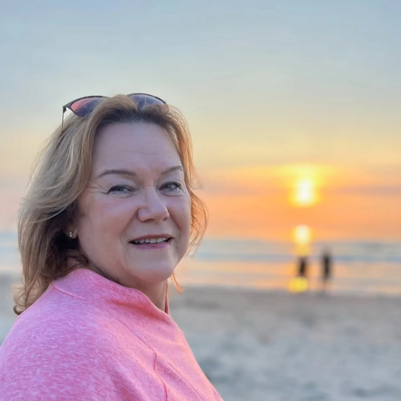 Jolene McDonough smiling on a sandy beach at sunset, with the sun setting over the ocean behind her